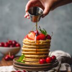 a stack of plant based pancakes topped with strawberries and cherries being drizzled with maple syrup
