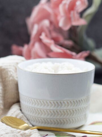 A white textured cup filled with creamy vegan rice pudding sits on a cloth next to a gold spoon, with pink flowers in the blurred background.