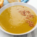 A bowl of smooth spicy butternut squash and sweet potato soup, garnished with crushed red pepper flakes, sits beside a slice of bread and a striped napkin in the background.