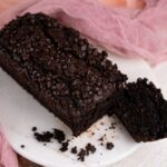 A loaf of vegan chocolate zucchini bread topped with chocolate chips sits on a round white platter, with one slice cut off and some crumbs scattered nearby.