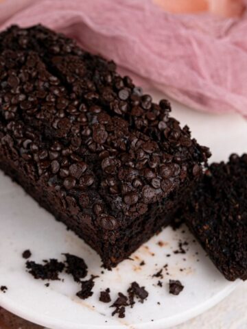 A loaf of vegan chocolate zucchini bread topped with chocolate chips sits on a round white platter, with one slice cut off and some crumbs scattered nearby.