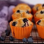 Vegan blueberry muffins in orange paper liners are arranged on a cooling rack, with fresh blueberries scattered nearby and a purple cloth in the background.
