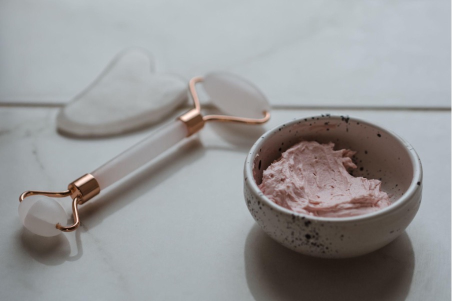 A facial roller, a gua sha tool, and a bowl of pink facial cream set on a white marble surface, showcasing an array of vegan skin treatments.