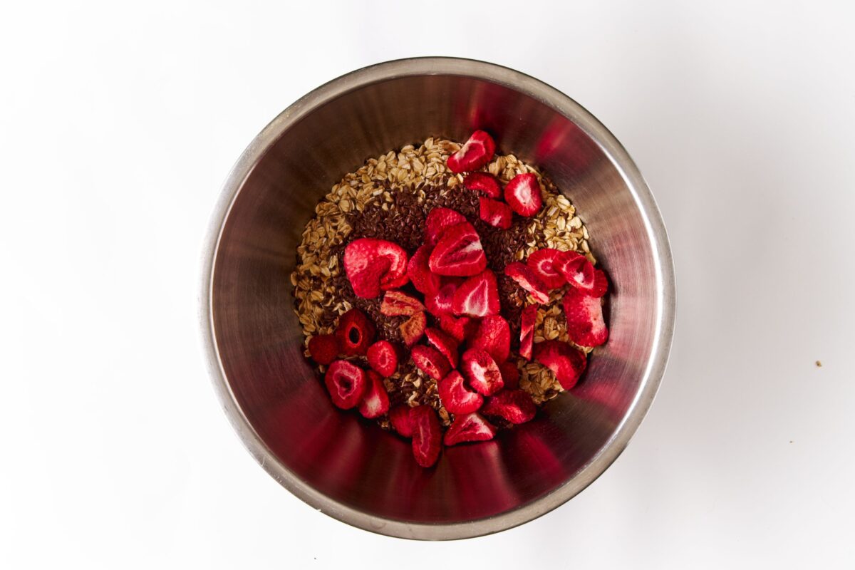 Vegan Strawberry and Cream Granola Protein Bars 219 A metal bowl containing oats, sliced freeze-dried strawberries, and other ingredients on a white surface.