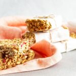 Strawberry protein bars wrapped in white paper and tied with string are placed on a peach-colored cloth, with unwrapped granola bars in the foreground.