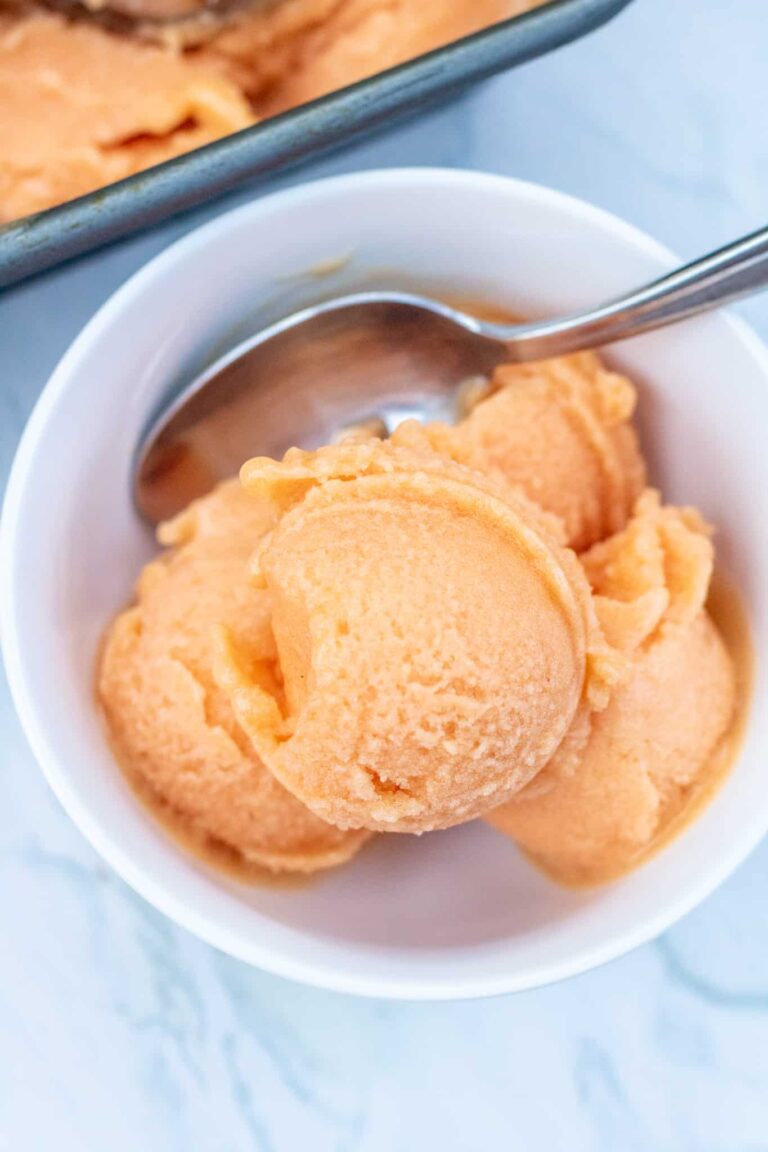 Easy Vegan Fruit Dessert Recipes You Will Love! 15 A white bowl contains three scoops of orange-colored sherbet with a metal spoon. A tray with more vegan fruit desserts is partially visible in the background.