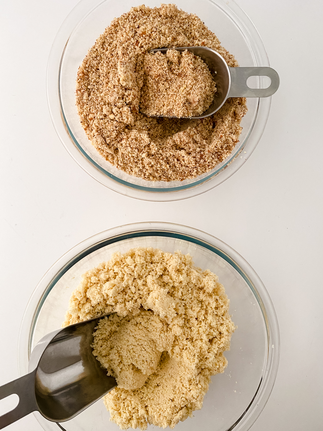 Two glass bowls contain different types of ground ingredients, each with a metal scoop. The top bowl holds a coarser brown mixture, while the bottom features a finer yellow blend. For those curious about how to make almond flour, it would resemble the fine texture seen in the bottom bowl.