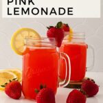 Two mason jar glasses filled with strawberry pink lemonade, garnished with lemon slices and strawberries, are placed on a white surface, surrounded by additional strawberries and a lemon slice.