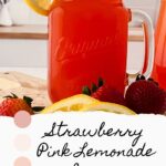 A mason jar filled with refreshing strawberry pink lemonade, garnished with a lemon slice and strawberries, sits on a wooden surface. A handwritten sign reads "Strawberry Pink Lemonade Recipe" in front.