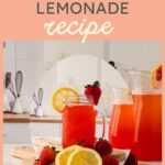 A mason jar filled with strawberry pink lemonade, garnished with lemon slices and strawberries, sits on a wooden cutting board next to a pitcher of the same refreshing drink. Lemons and strawberries are scattered nearby.