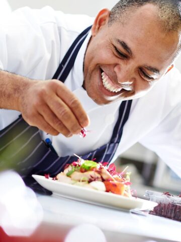 A smiling chef in a white coat and striped apron is garnishing a dish with herbs in a kitchen, showcasing delicious vegan menu ideas.
