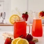 A glass mason jar and a pitcher filled with the best homemade strawberry pink lemonade recipe are placed on a kitchen counter. The jar and pitcher are garnished with lemon slices and strawberries, surrounded by additional fruit.