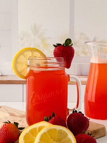 A glass mason jar and a pitcher filled with the best homemade strawberry pink lemonade recipe are placed on a kitchen counter. The jar and pitcher are garnished with lemon slices and strawberries, surrounded by additional fruit.