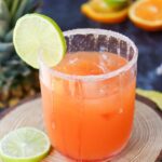 A glass filled with an orange-colored drink, garnished with a lime slice on the rim, sits on a wooden coaster. Another lime slice and orange slices are in the blurred background.