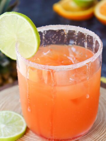 A glass filled with an orange-colored drink, garnished with a lime slice on the rim, sits on a wooden coaster. Another lime slice and orange slices are in the blurred background.