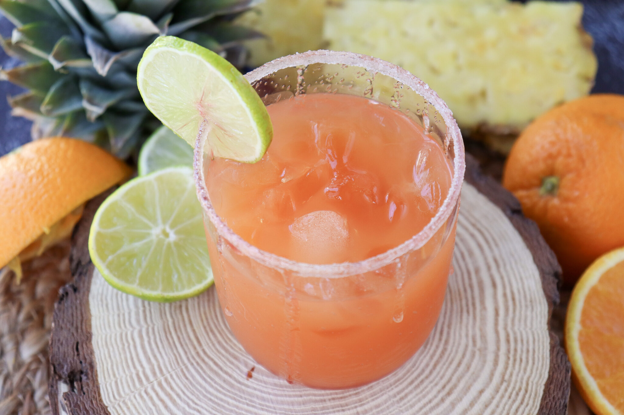A glass of orange and lime cocktail with ice, garnished with a lime slice, sits on a wooden surface surrounded by fresh orange and lime slices.