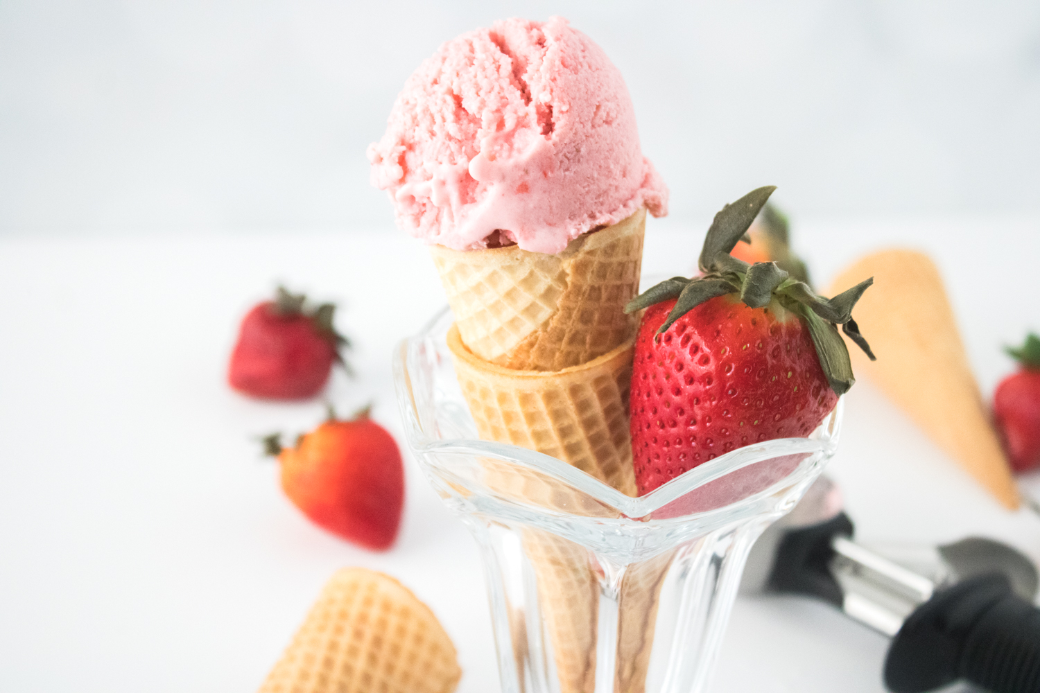 A waffle cone with a scoop of strawberry ice cream is placed in a glass, accompanied by a fresh strawberry. Other strawberries and an ice cream scoop are visible in the background.
