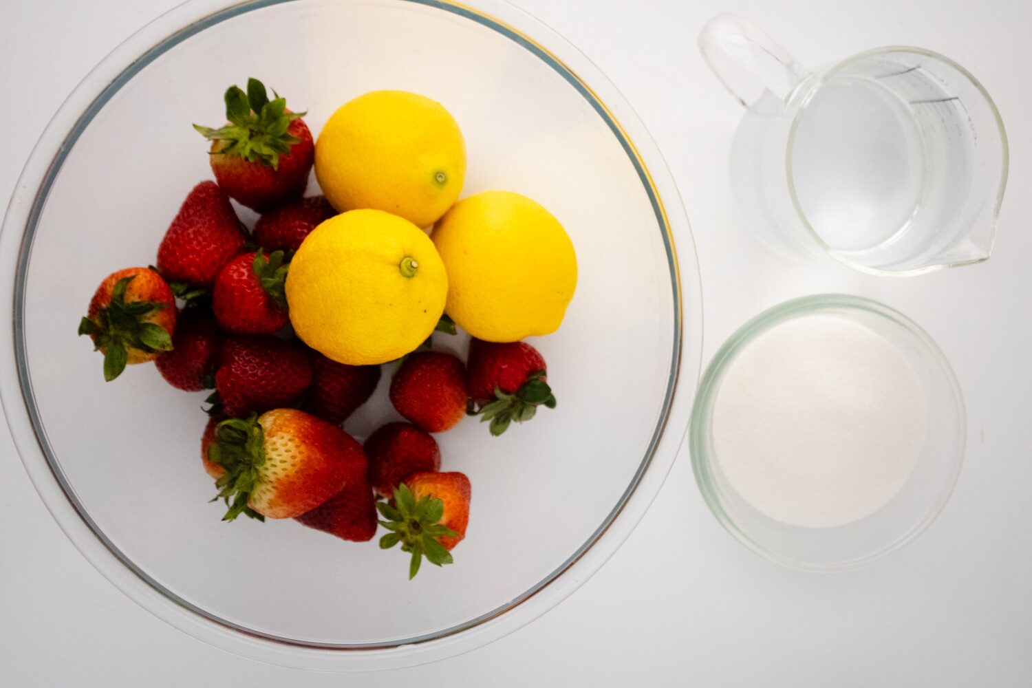 Best Homemade Strawberry Pink Lemonade Recipe 170 A bowl of lemons and strawberries, a bowl of sugar, and a measuring cup of water on a white surface.