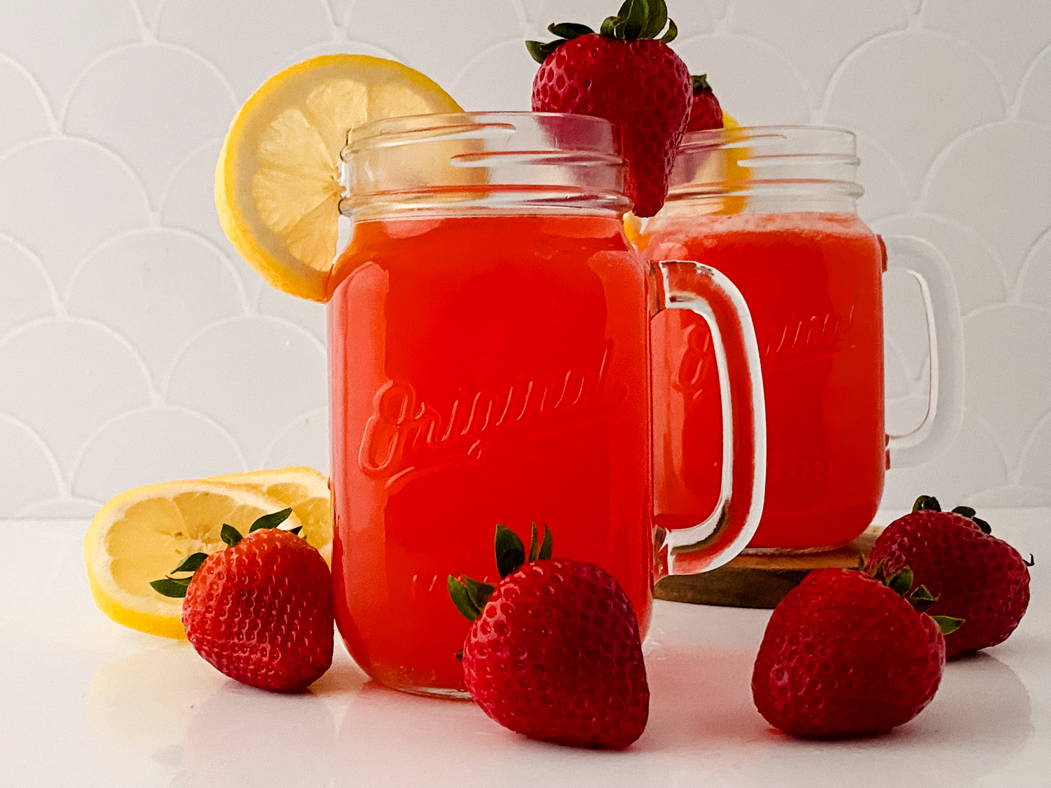 Two mason jars filled with a red drink, garnished with lemon slices and strawberries, set on a white surface with additional strawberries and lemon slices around.