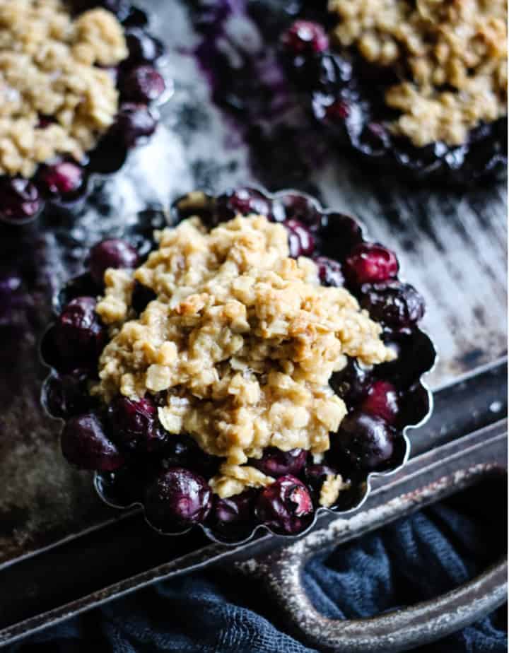 30 Delicious Vegan Blueberry Dessert Recipes 9 Three individual vegan blueberry crisps served in small baking dishes on a dark background.