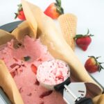 A pan of vegan strawberry ice cream with chunks of strawberries, partially scooped with an ice cream scooper. An empty waffle cone and whole strawberries are placed in the background.