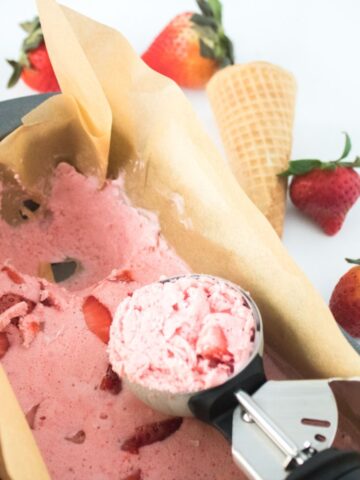 A pan of vegan strawberry ice cream with chunks of strawberries, partially scooped with an ice cream scooper. An empty waffle cone and whole strawberries are placed in the background.