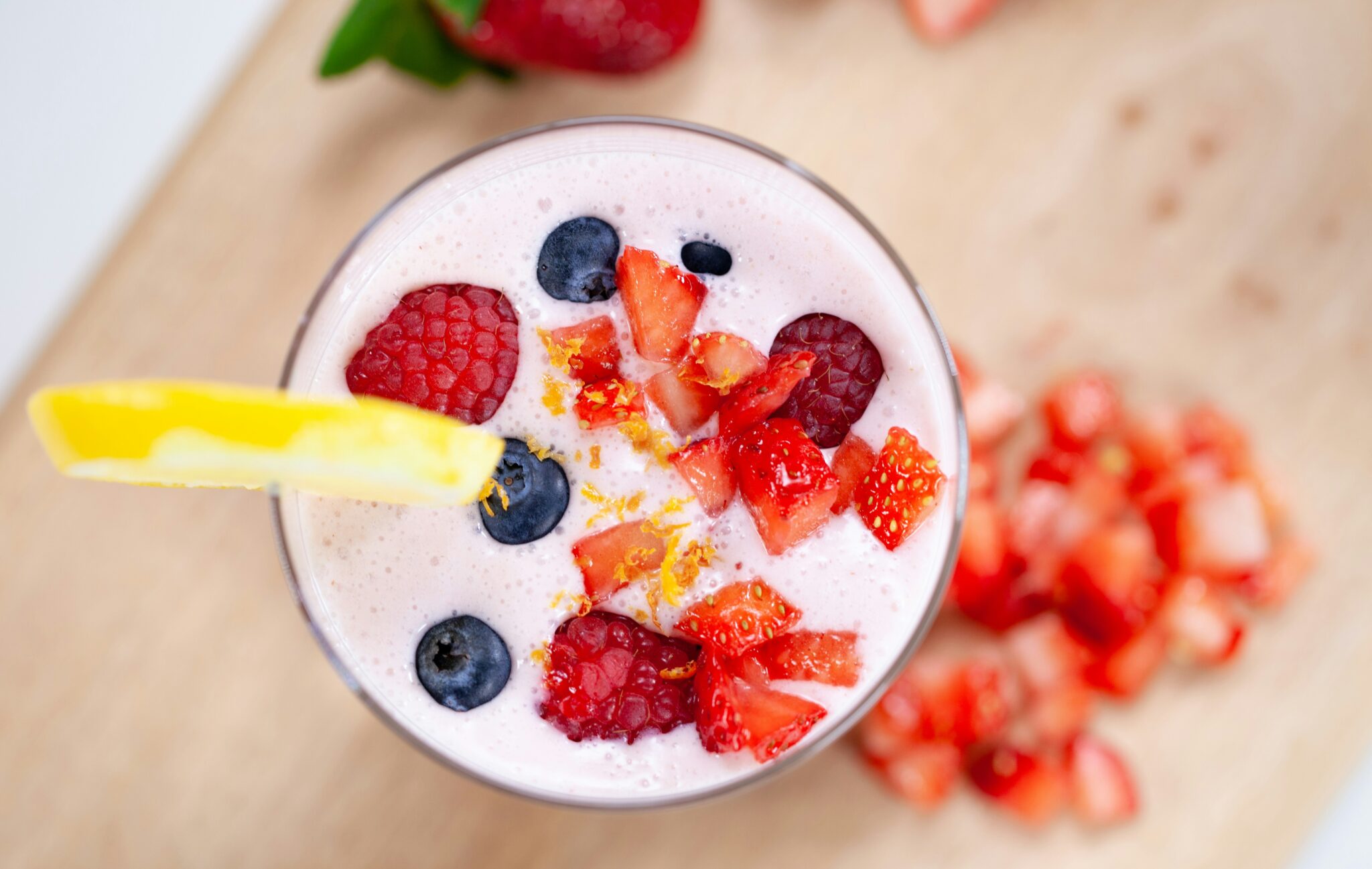Top view of a smoothie in a glass, garnished with raspberries, blueberries, strawberry pieces, lemon zest, and a lemon slice—an ideal treat for those learning how to transition to a plant-based diet. Chopped strawberries adorn the background.