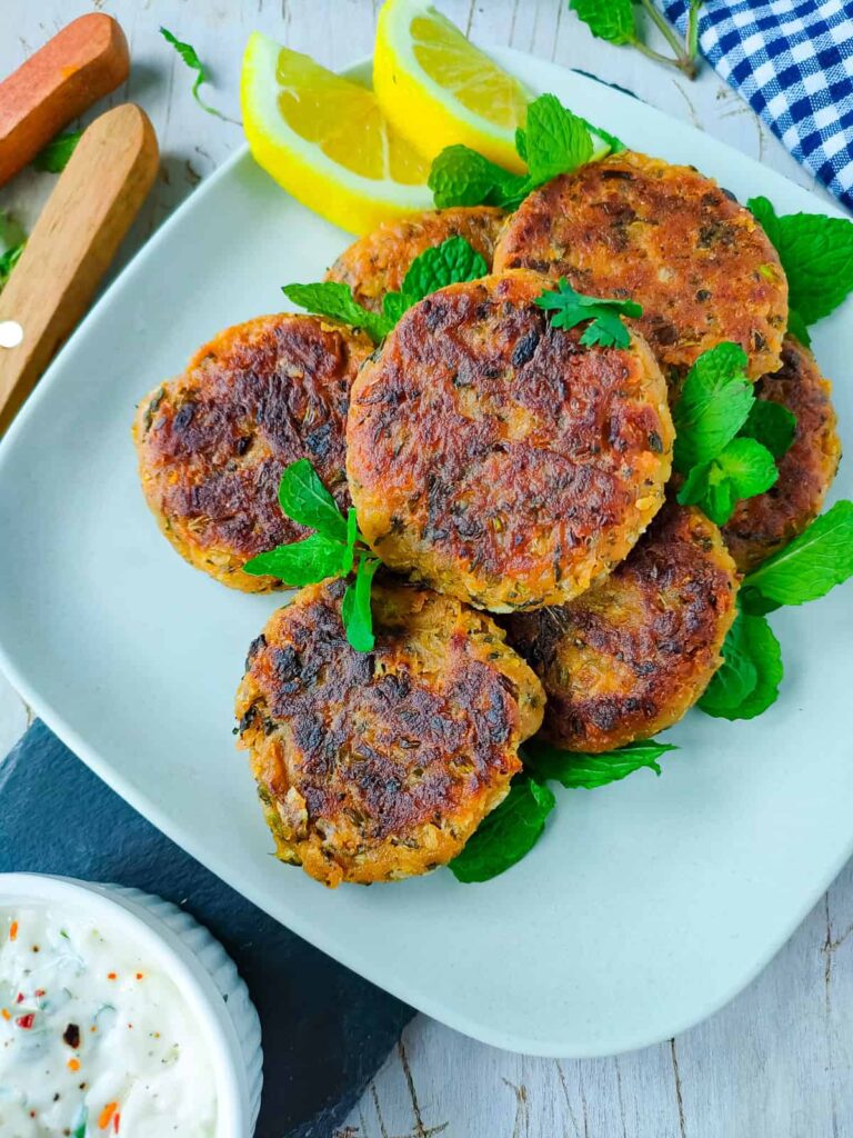 The Best High Protein Plant Based Snacks For On-The-Go 29 A plate of plant-based snacks, perfectly fried patties garnished with lemon slices and green herbs. A high-protein dipping sauce is placed nearby.