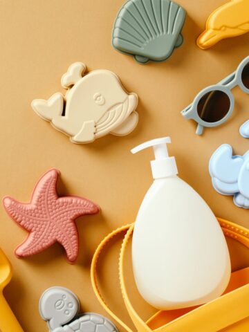 Silicone vegan beach toys, including a shovel, starfish, whale, shell, and crab shapes, sunglasses, a sunscreen bottle, and a yellow strap are laid out on a brown background.
