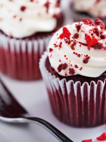 Close-up of red velvet cupcakes topped with cream cheese frosting, red velvet crumbs, and heart-shaped sprinkles made vibrant with Red 40. A fork lies on the side.