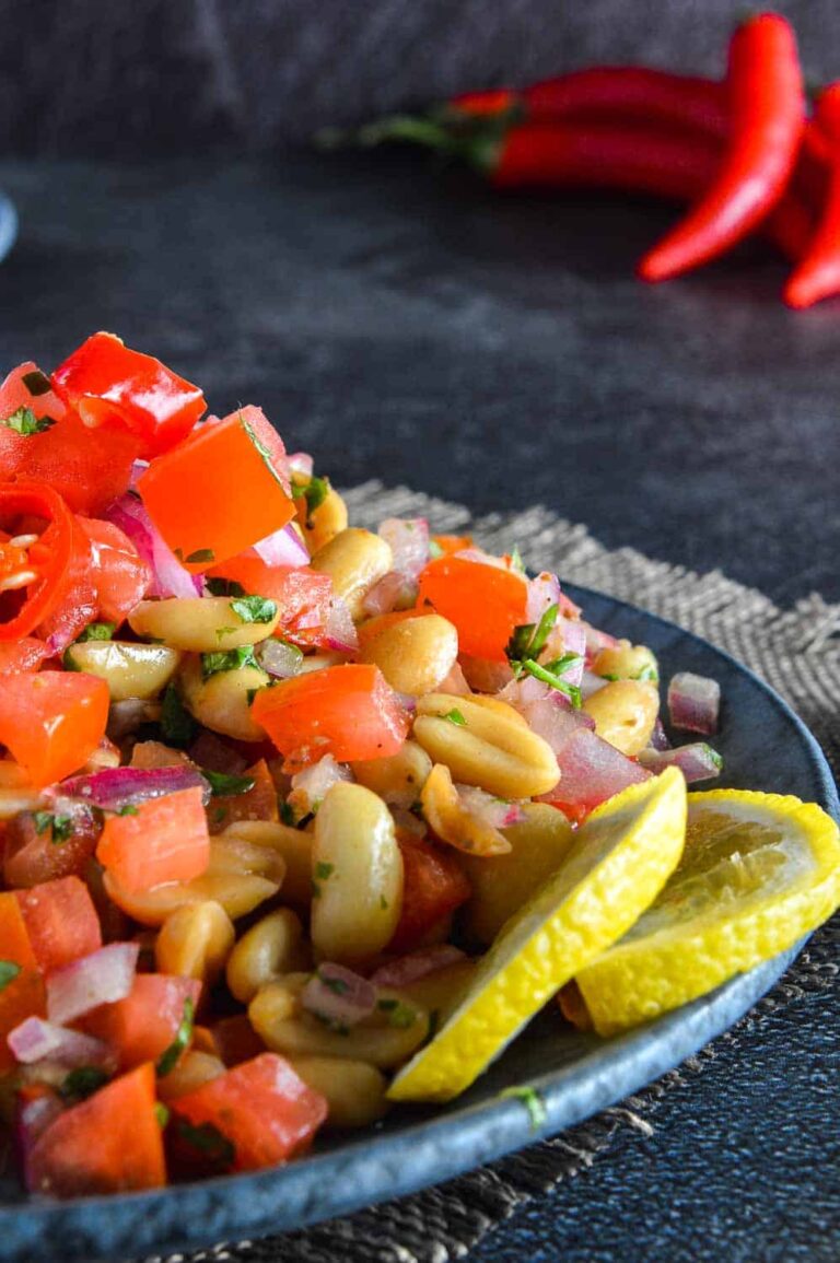The Best High Protein Plant Based Snacks For On-The-Go 24 A plate of high-protein chopped tomatoes and beans garnished with lemon slices on a textured surface, with red chili peppers in the background.