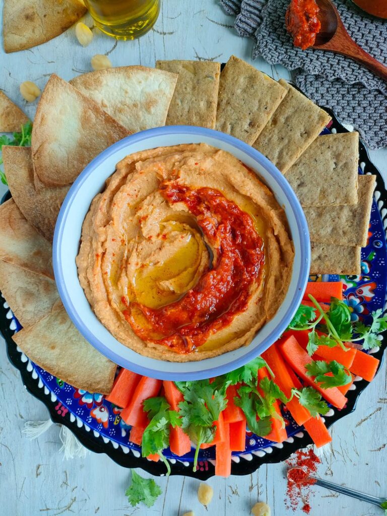 The Best High Protein Plant Based Snacks For On-The-Go 30 A plate of hummus topped with red pepper sauce, surrounded by pita bread, carrot sticks, and fresh cilantro on a decorated round tray-perfect plant-based snacks for an on-the-go lifestyle.