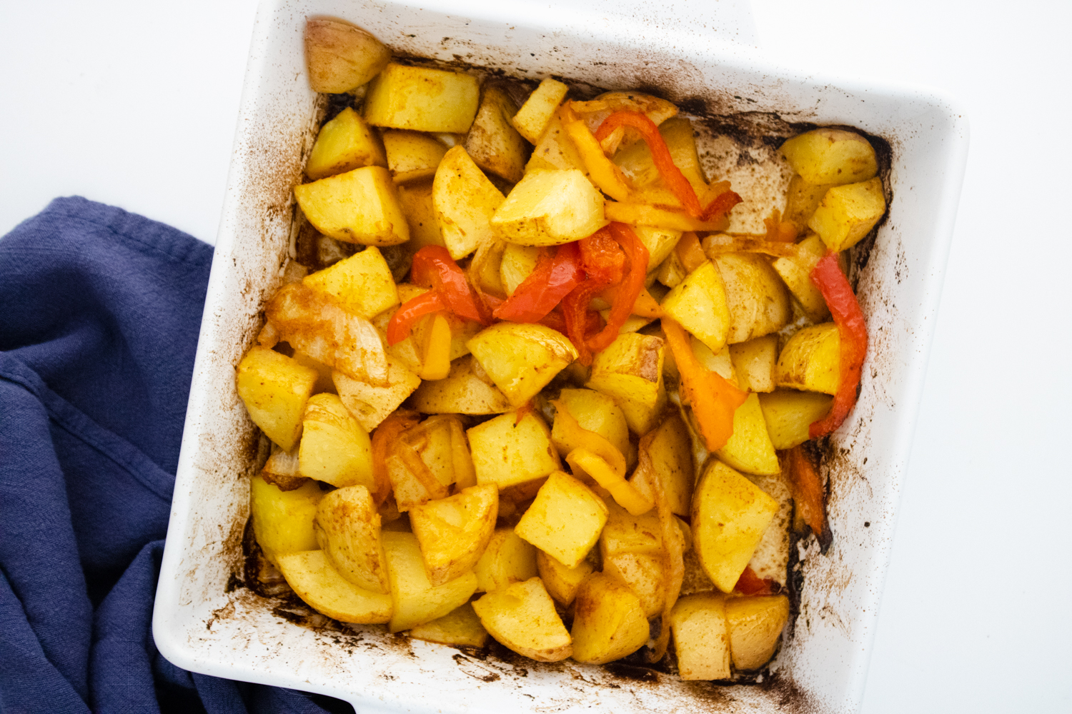 A white baking dish filled with roasted potato and bell pepper chunks. The dish sits on a white surface next to a blue cloth.