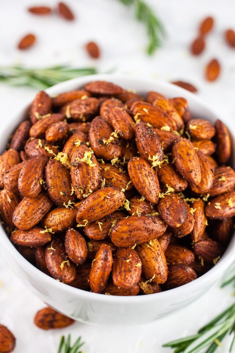 The Best High Protein Plant Based Snacks For On-The-Go 9 A bowl filled with seasoned roasted almonds, garnished with lemon zest, rests amidst scattered almonds and sprigs of rosemary. This high-protein, plant-based snack is perfect for those seeking a nutritious boost.