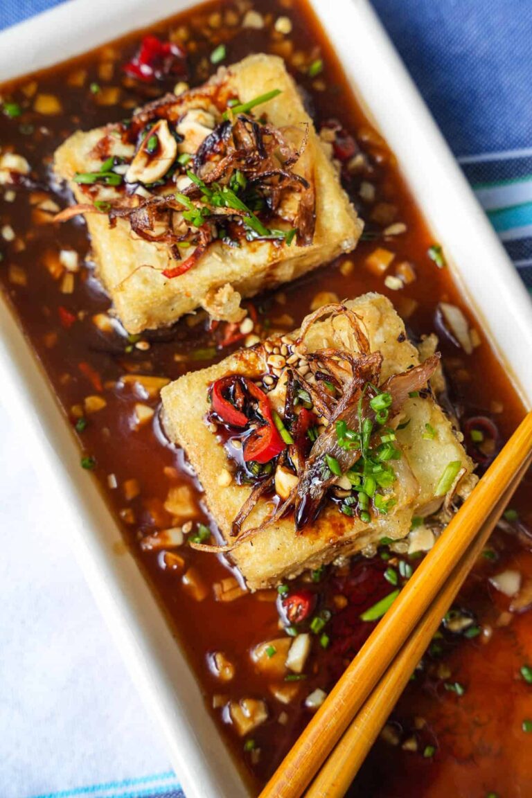 The Best High Protein Plant Based Snacks For On-The-Go 17 A rectangular plate with two pieces of high-protein fried tofu topped with chopped chiles, nuts, and herbs, served in a soy-based sauce. Yellow chopsticks are placed beside the tofu for a delightful plant-based snack.