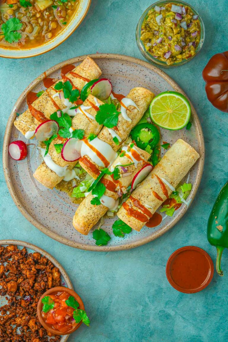 The Best High Protein Plant Based Snacks For On-The-Go 16 A plate with five rolled tacos drizzled with sauces, garnished with radish slices and cilantro, accompanied by a lime half and jalapeño slices. Surrounding dishes contain various toppings and plant-based snacks for an on-the-go meal.