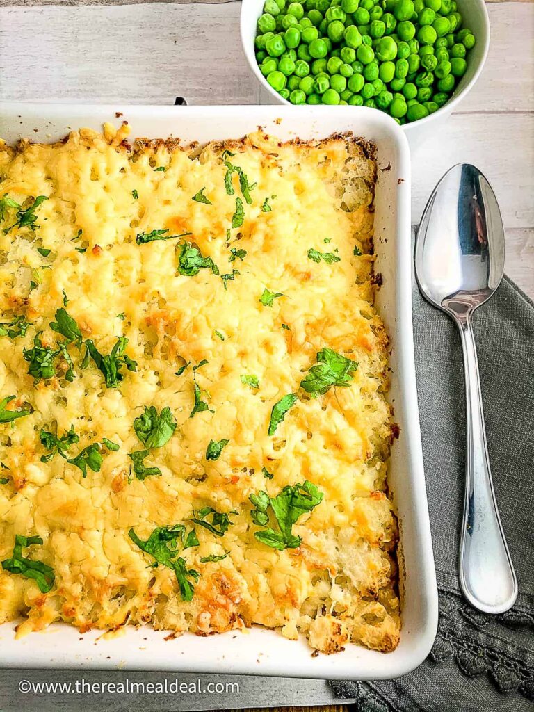 High Calorie Vegan Recipes - Meals For Weight Gain 18 A high-calorie vegan casserole, topped with melted cheese and fresh herbs, sits invitingly in a white dish beside a bowl of green peas. A spoon rests on a grey napkin, completing the cozy table scene.