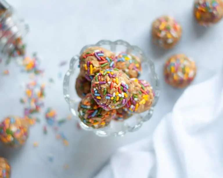The Best High Protein Plant Based Snacks For On-The-Go 18 A glass bowl filled with sprinkle-covered, high-protein cookie dough bites is shown from above, surrounded by more cookie dough bites and scattered sprinkles on a white surface.