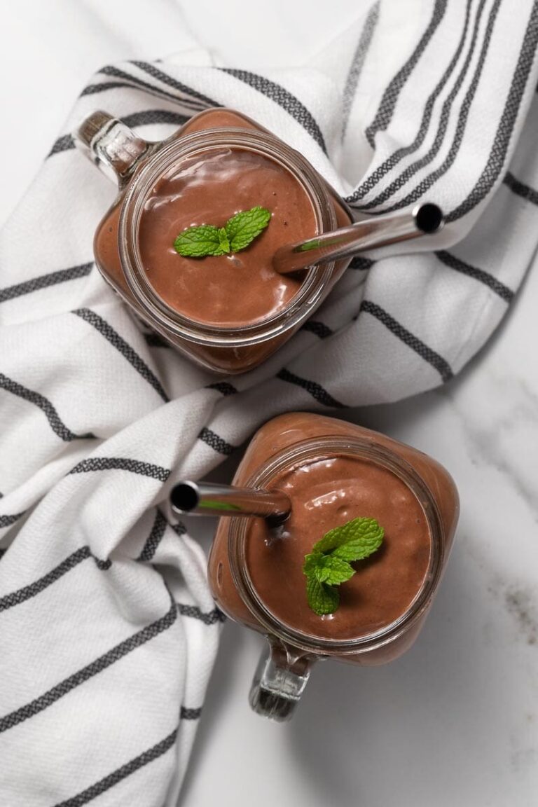 Two glasses of rich, chocolate smoothies with stainless steel straws, garnished with mint leaves, rest invitingly on a striped cloth. Perfect for those exploring high-calorie vegan recipes.