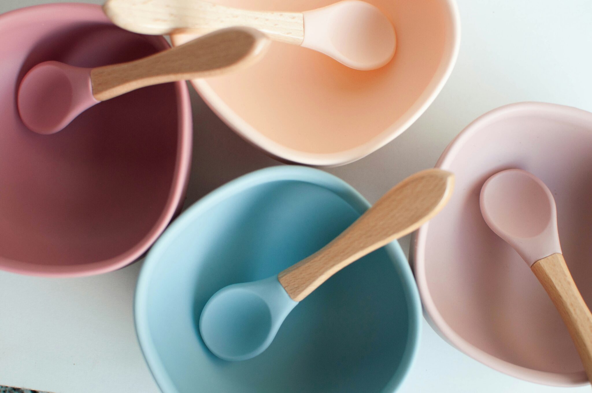 Four silicone bowls in pink, peach, and blue with matching wooden-handled spoons are arranged closely together on a white surface. These vibrant bowls demonstrate that silicone is vegan-friendly and stylishly practical for modern kitchens.