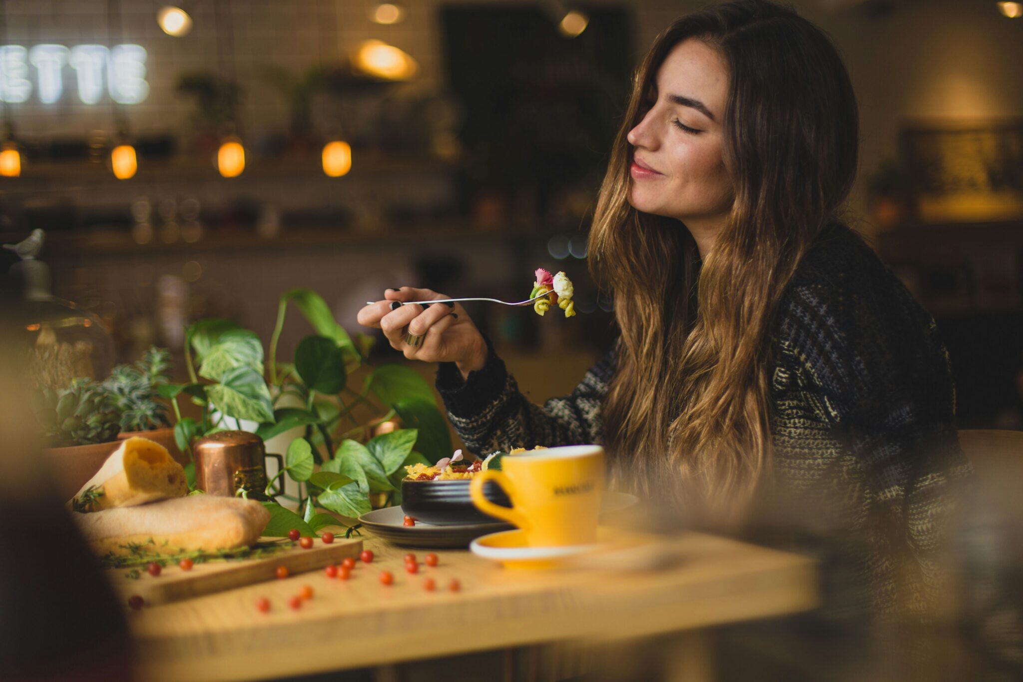 A woman savors her meal from a fork at the restaurant table, surrounded by high-calorie vegan recipes. A cup and various dishes adorn the table, with lush indoor plants providing a serene backdrop.