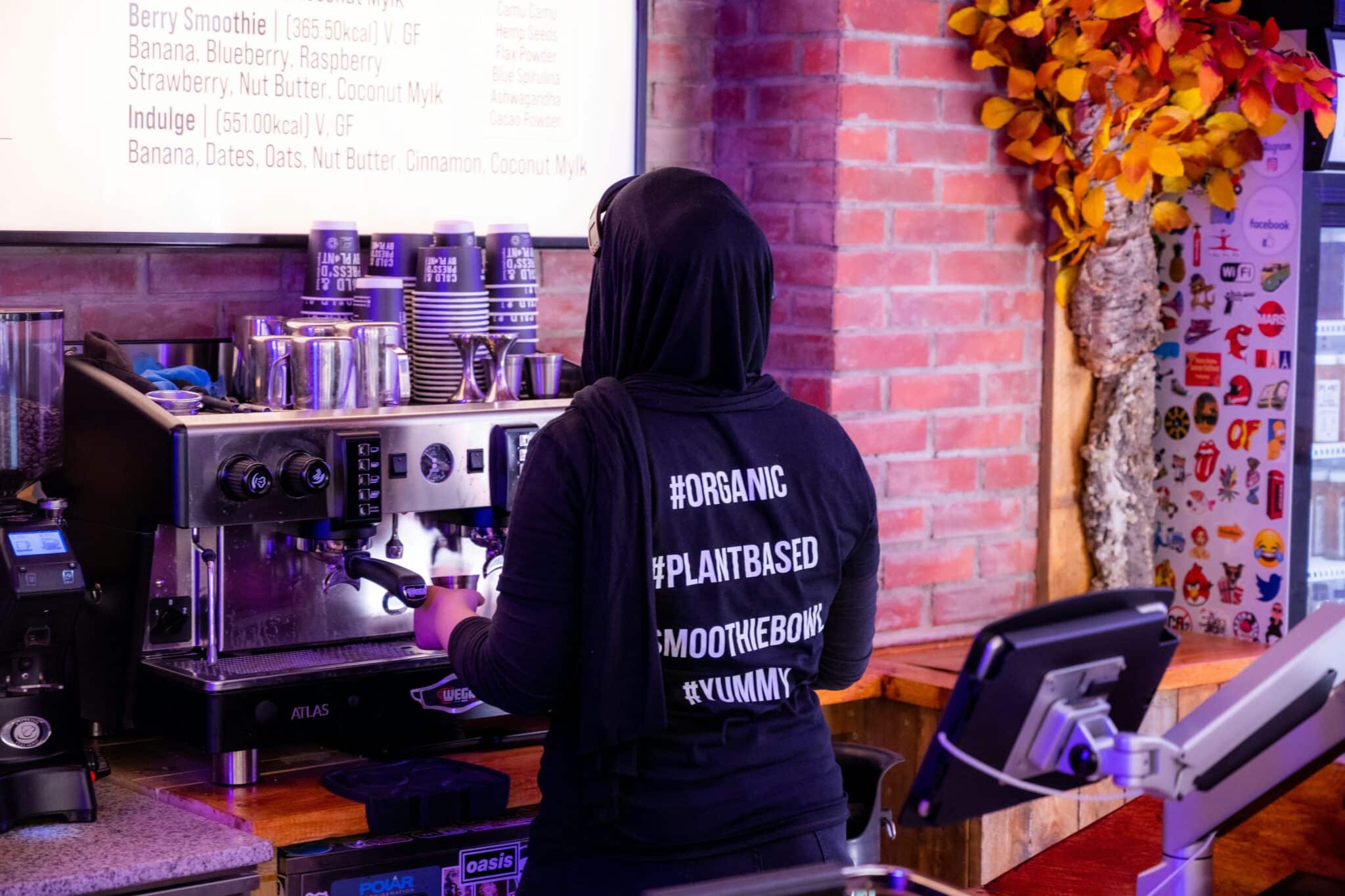 A person in a black sweatshirt adorned with hashtags operates a coffee machine in a café, perhaps serving up plant-based options for those curious about what "plant-based" means.
