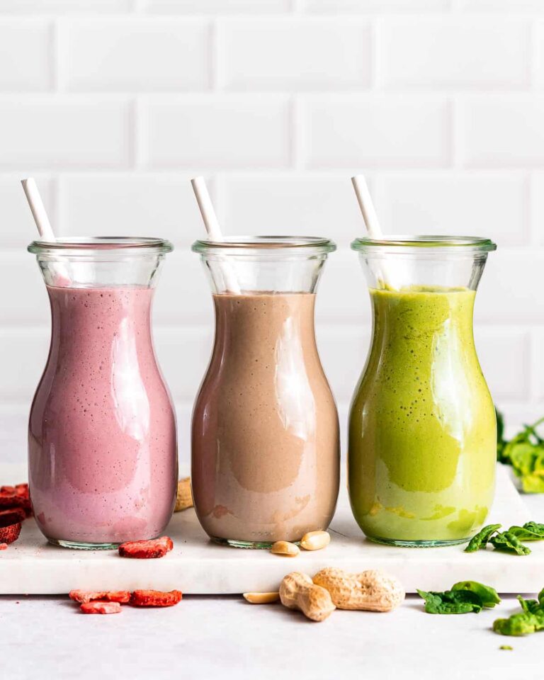 The Best High Protein Plant Based Snacks For On-The-Go 27 Three glass bottles with straws, filled with pink, brown, and green smoothies respectively, are arranged on a countertop with scattered high-protein snacks like peanuts and dried fruit nearby.