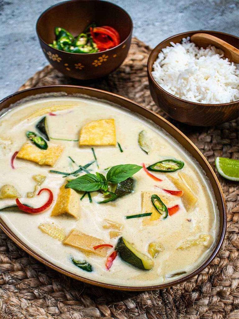High Calorie Vegan Recipes - Meals For Weight Gain 8 A bowl of high-calorie Thai green curry with tofu, vegetables, and basil leaves sits alongside white rice and sliced chili peppers on a woven mat.