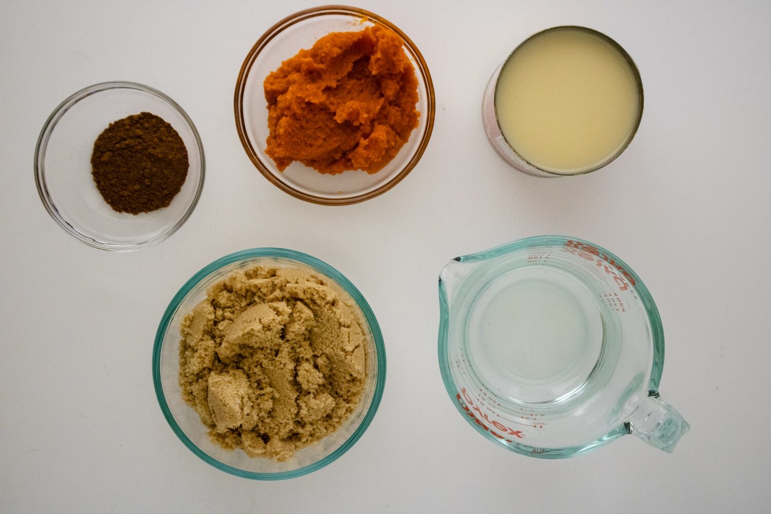 Homemade Pumpkin Spice Simple Syrup Recipe 109 Overhead view of pumpkin spice simple syrup ingredients including pumpkin puree, vegan condensed milk, brown sugar, water and spices arranged on a white surface.