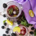 A pitcher and glass of blackberry mojito with mint and ice, garnished with lime slices. Blackberries, a lime slice, and mint leaves are scattered on a marble surface with a purple fabric background.