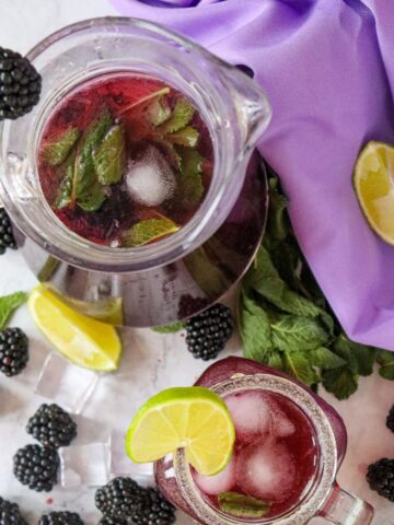 A pitcher and glass of blackberry mojito with mint and ice, garnished with lime slices. Blackberries, a lime slice, and mint leaves are scattered on a marble surface with a purple fabric background.