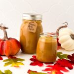 Two jars of pumpkin spice syrup stand amid autumn leaves, flanked by small decorative pumpkins. One jar has a labeled tag reading "Pumpkin Spice Syrup." The background is a white tiled wall.
