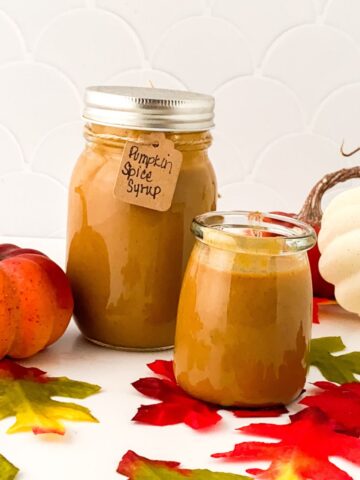 Two jars of pumpkin spice syrup stand amid autumn leaves, flanked by small decorative pumpkins. One jar has a labeled tag reading "Pumpkin Spice Syrup." The background is a white tiled wall.
