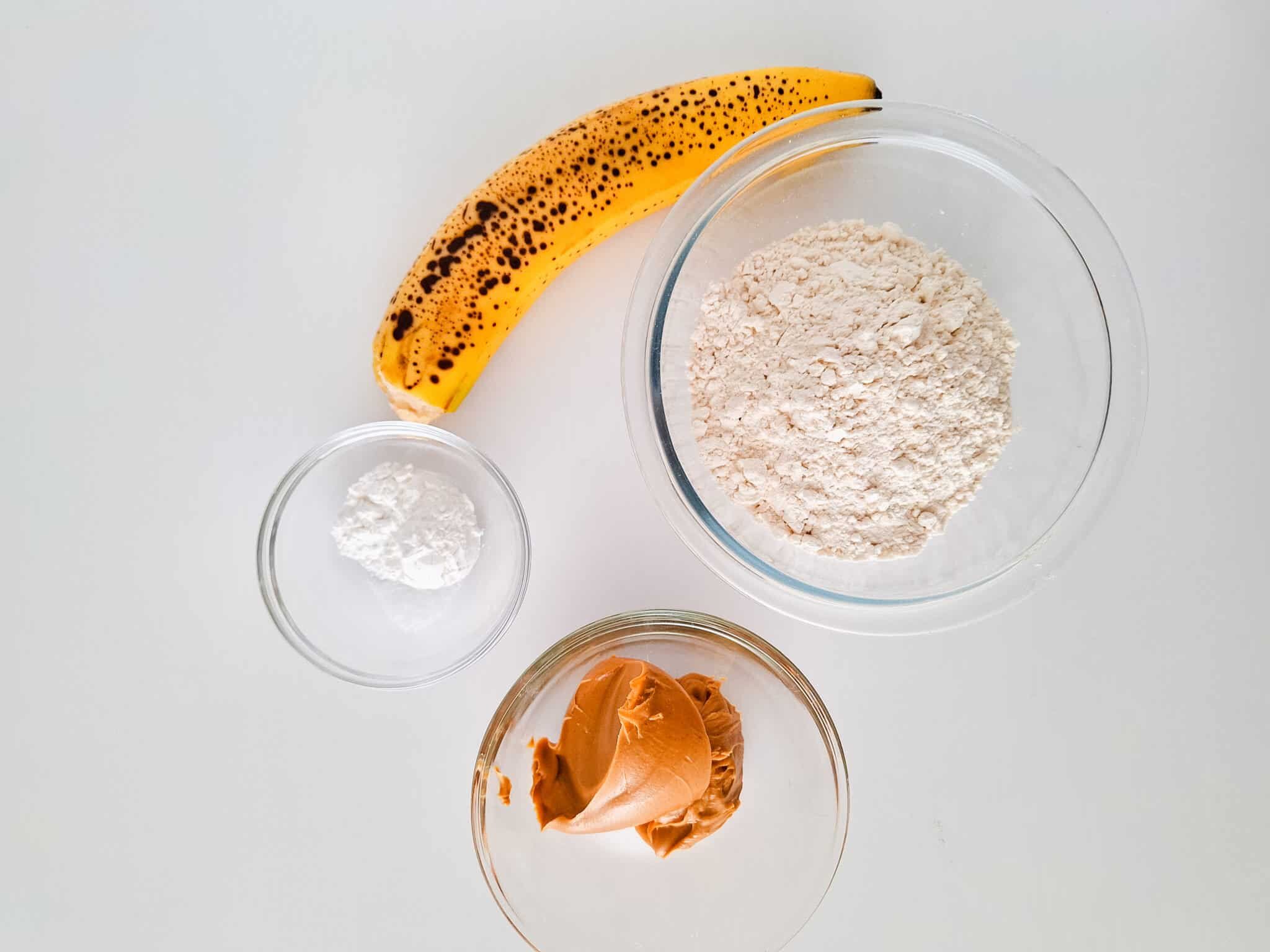 Vegan Banana Peanut Butter Cookies For Babies 83 Ripe banana, bowls of flour, peanut butter, and baking powder on a white surface.